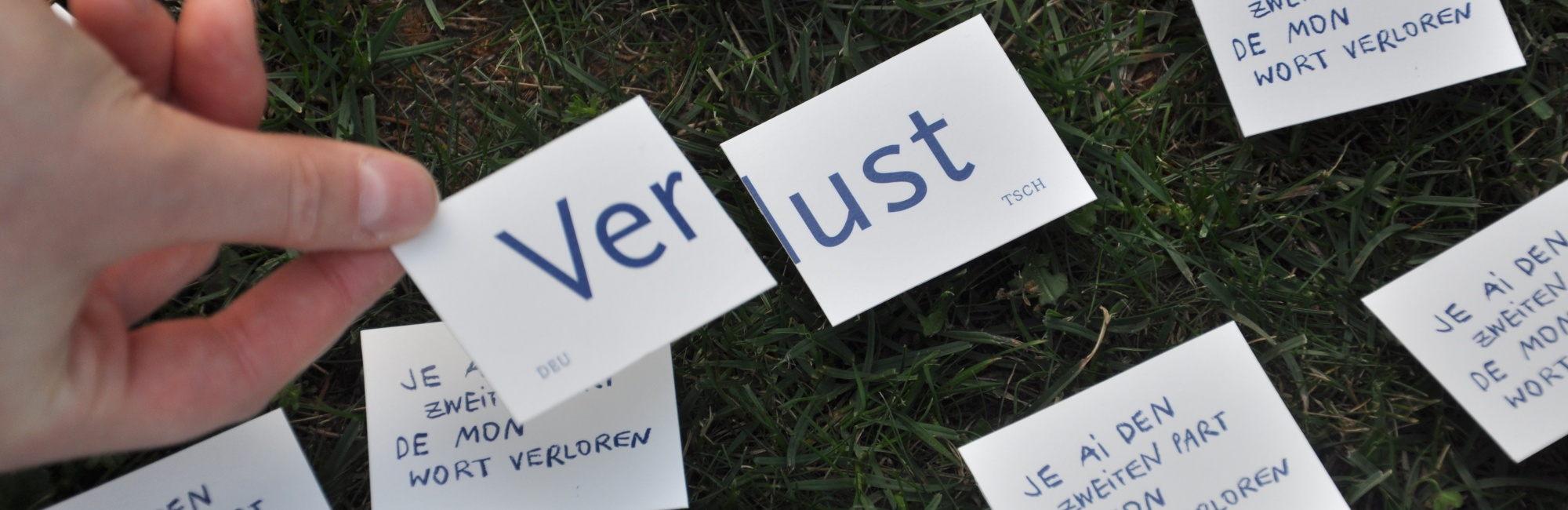 Auf einer Wiese sind Papierschnipsel hingelegt, die Schnipsel Ver und lust stehen hierbei im Vordergrund und werden von einer Hand gehalten