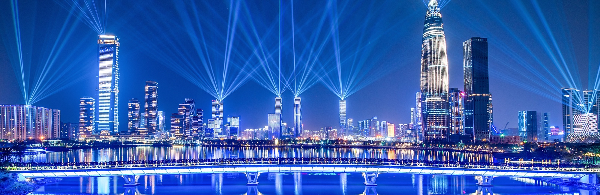 Skyline einer Stadt in der Dunkelheit zu sehen, im Vordergrund spiegelt sich die Skyline im Wasser des Hafens, von den Hochhäusern aus leuchten die Strahlen von Scheinwerfern in den Himmel