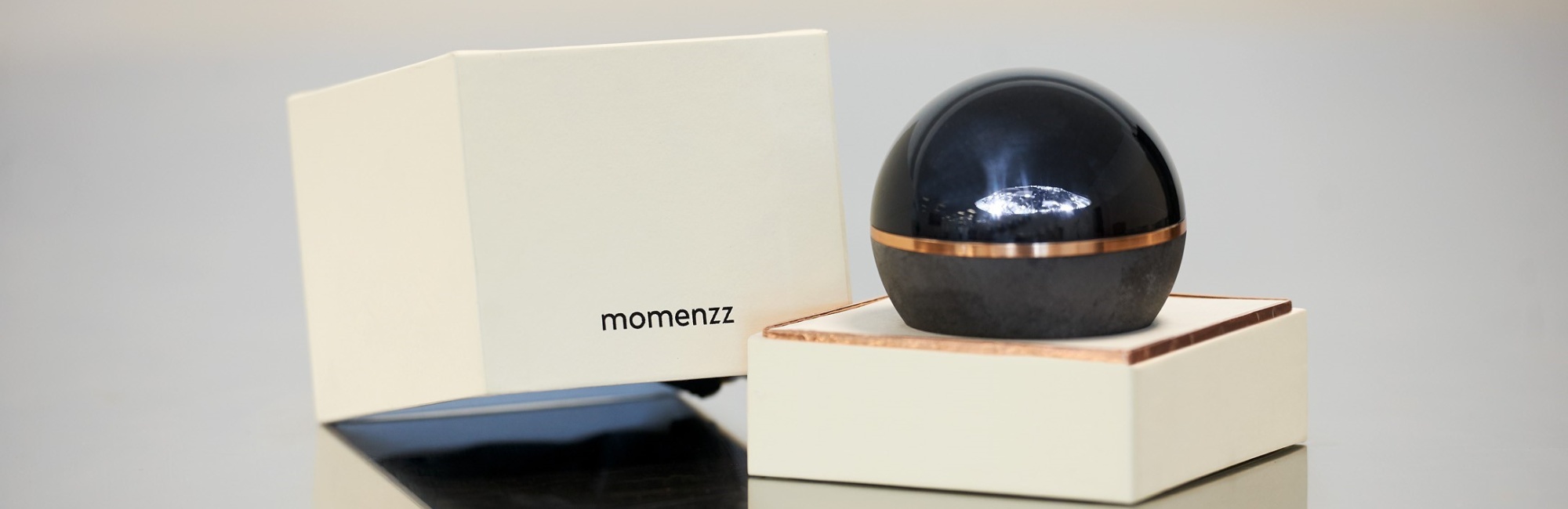 auf einem Spiegelunergrund steht eine weiße Box die geöffnet ist, auf dem unteren Teil der Box steht eine schwarze Kugel aus Glas mit einem goldenen horizontalen Strich, der Deckel der Box liegt links daneben und es befindet sich der Schriftzug "momenzz" darauf