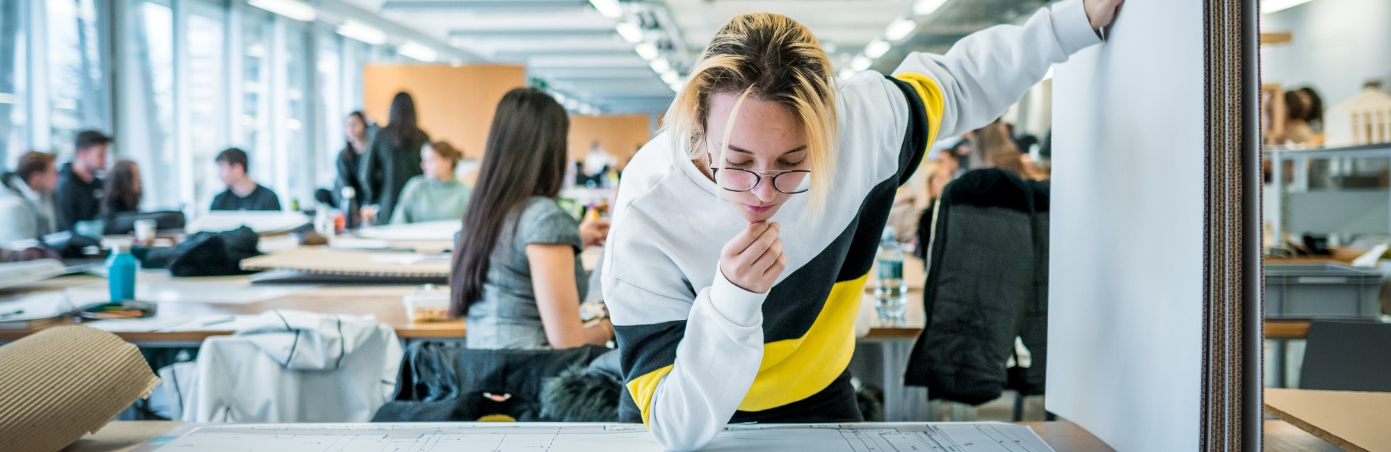 Studierende betrachtet eine Skizze die auf einem Tisch liegt, im Großraumbüro