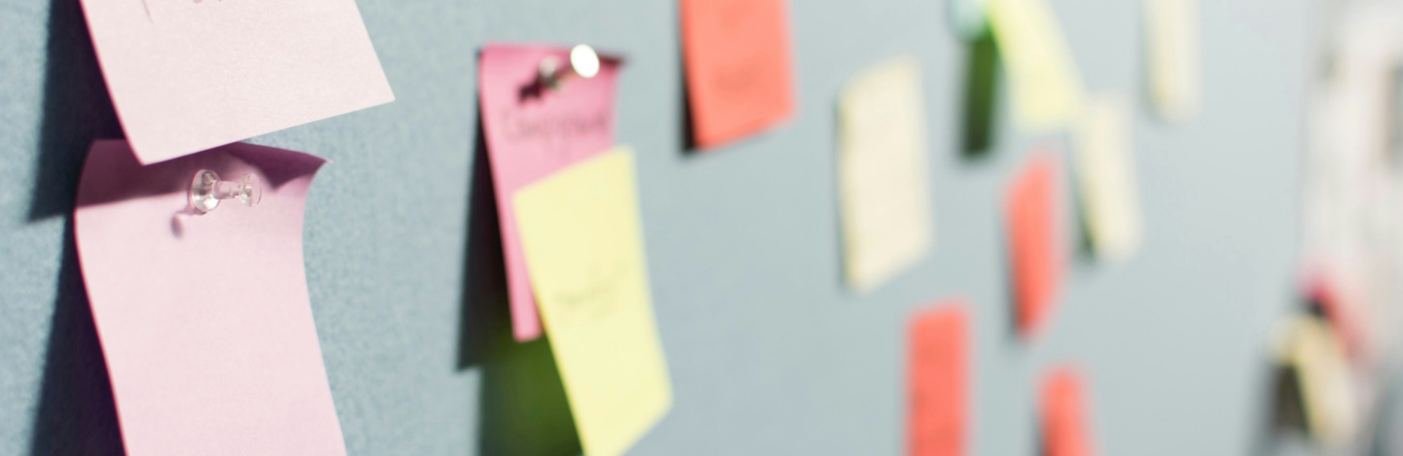 Das Bild zeigt eine graue Pinnwand auf der pastellfarbene Post-Its stecken mit Pins
