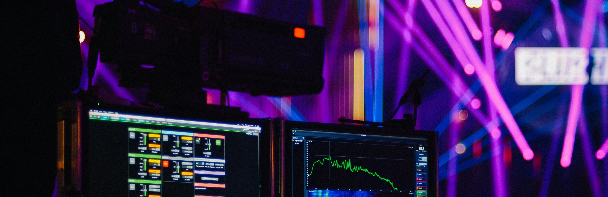 Im dunklen Hintergrund sieht man diverse bunte Lichtstrahlen von Schinwerfern, im Vordergrund kann man Eventtechnik-Monitore sehen mit Lichttechnik und Audioprogrammen