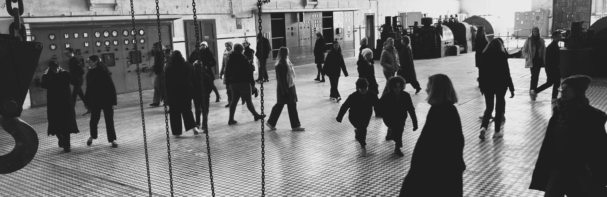 Schwarz-Wei&szlig;-Bild das eine Maschinenhalle darstellt, &uuml;ber den Boden gehen unterschiedliche Menschen kreuz und quer, alle dunkel gekleidet