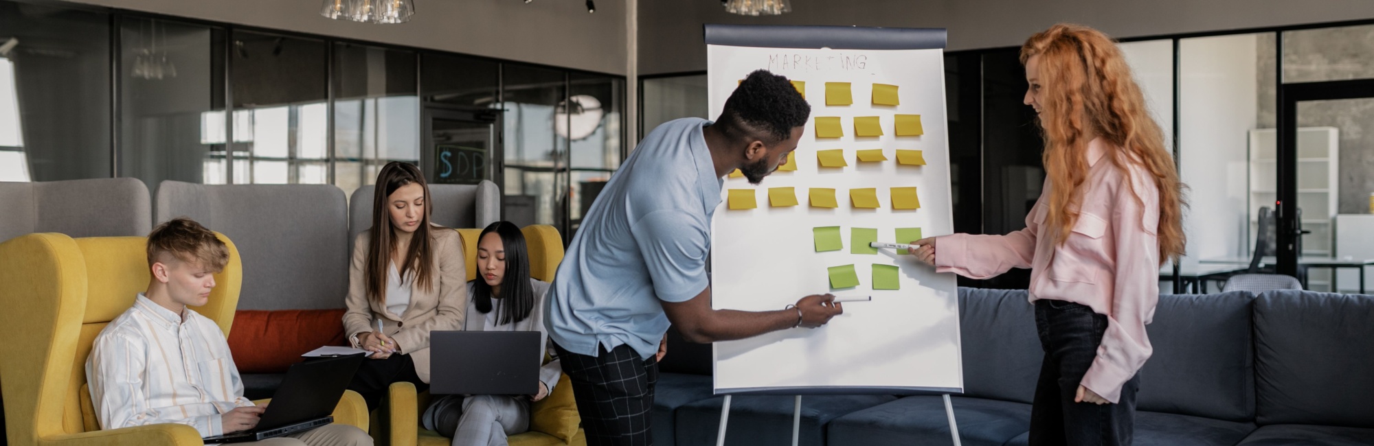 Zwei Studenten an einem Flipchart mit Post-Its. Im Hintergrund sitzen drei weitere Studenten mit Laptops.