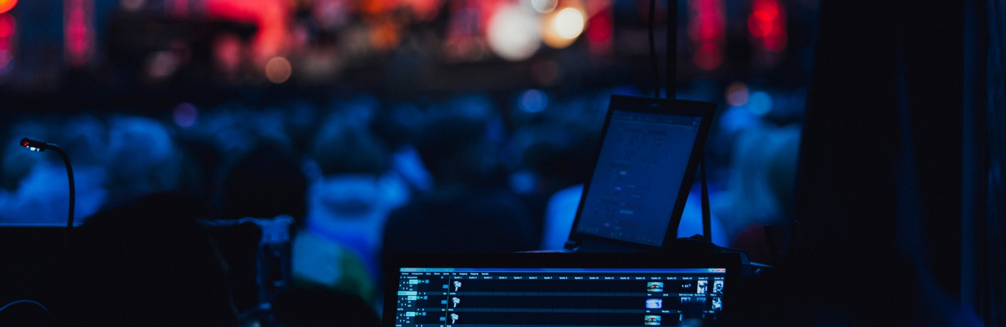 Eventhalle während eines Events zu sehen, die Lichtstimmung ist rot und blau und sehr dunkel gehalten, im Vordergrund ist ein Audiomischpult zu sehen mit weiteren technischen Geräten